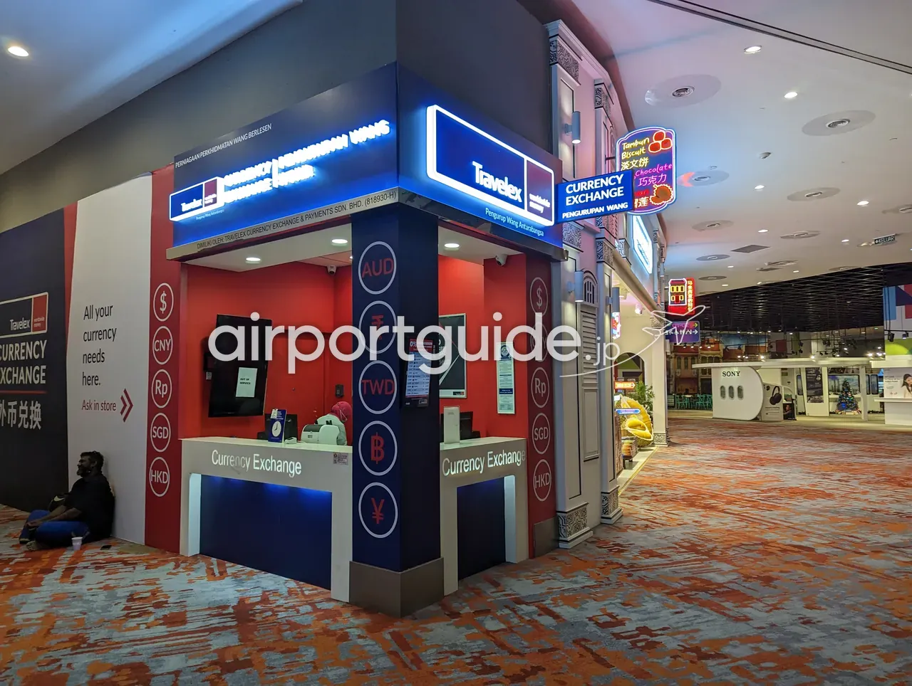 KLIA制限エリア内、絨毯敷きのショッピングストリートに立つ赤・白・紺のTravelex両替ブース。「Travelex CURRENCY EXCHANGE」「Pengurupan Wang」「外币兑换」「All your currency needs here」「0% Commission」の表示、柱にAUD/CNY/Rp/TWD/฿/HKDなどの通貨コード。右手にヘリテージ調の店構えの土産店とSonyショップ。