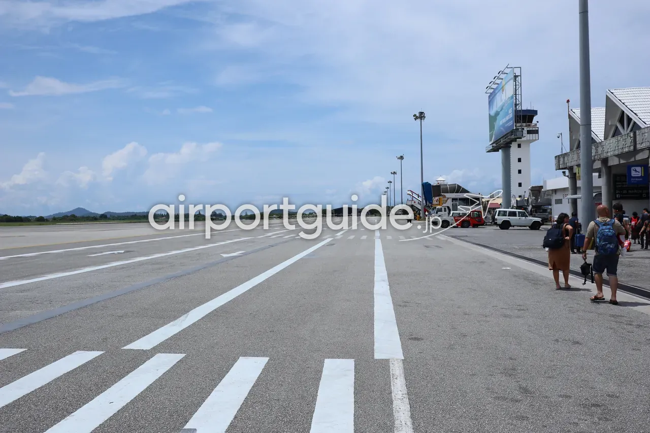 ランカウイ国際空港の広く開けた駐機場を、徒歩でターミナルへ向かう眺め。青空の下に白い誘導線が延び、右奥に白い管制塔と三角屋根のターミナル、ビルボード、トーイングカー。