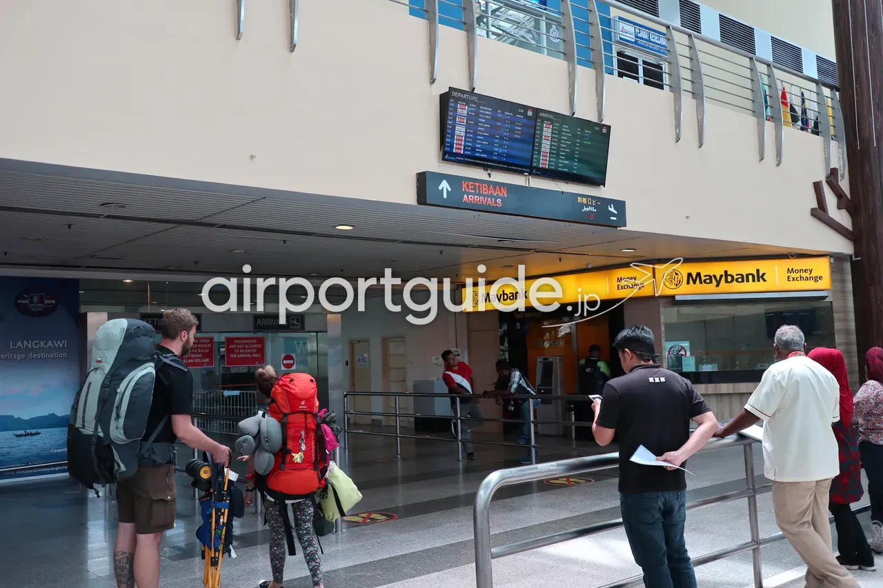 ランカウイ国際空港の到着ホール。頭上に『KETIBAAN ARRIVALS 到着』の多言語案内板とフライト情報モニター、黄色い『Maybank Money Exchange』の両替カウンター、『LANGKAWI heritage destination』のポスター、大きな荷物を背負ったバックパッカーの旅行者。