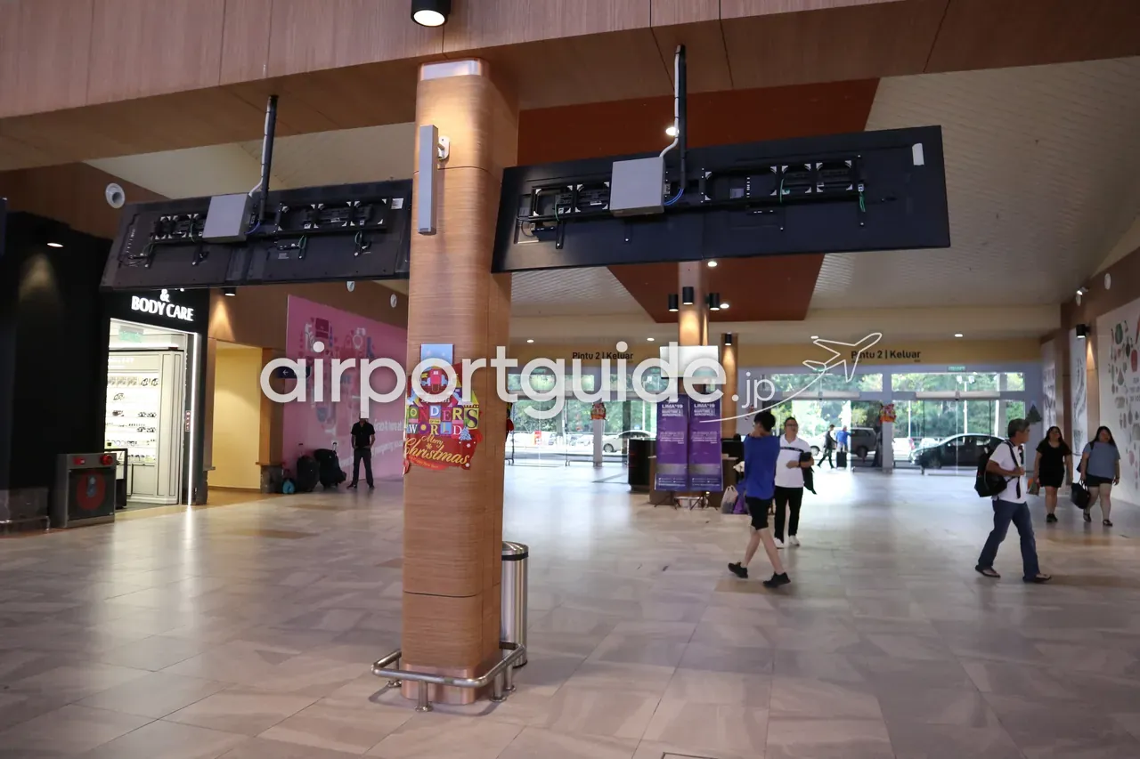 ランカウイ国際空港のコンコース内部。木目調パネルの天井と太い木目の柱、右手に『Pintu 2 | Keluar』（出口2番）の案内、左手に化粧品の売店、明るいガラス出口へ向かって歩く旅客。