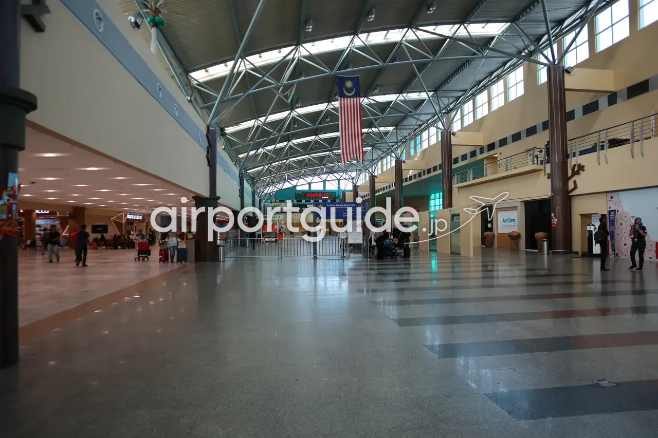 ランカウイ国際空港のメインホール。鉄骨トラスの採光天窓の下にマレーシア国旗が吊られ、奥に『KASTAM DIRAJA MALAYSIA』（税関）ゲートと『AeroDarat』のカウンター、まだら模様の石の床。