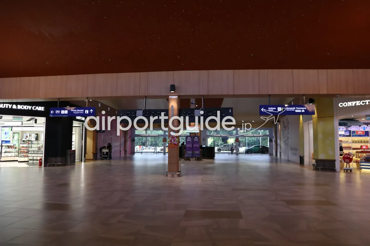 ランカウイ国際空港の到着・送迎エリア。星型ライトを散らした暗いブラウン調の天井、『Ketibaan Arrivals』『Mengambil Penumpang Pick-up Point』の青い案内板、ガラス出口の外に駐車する車、右手にコンビニ的な売店。