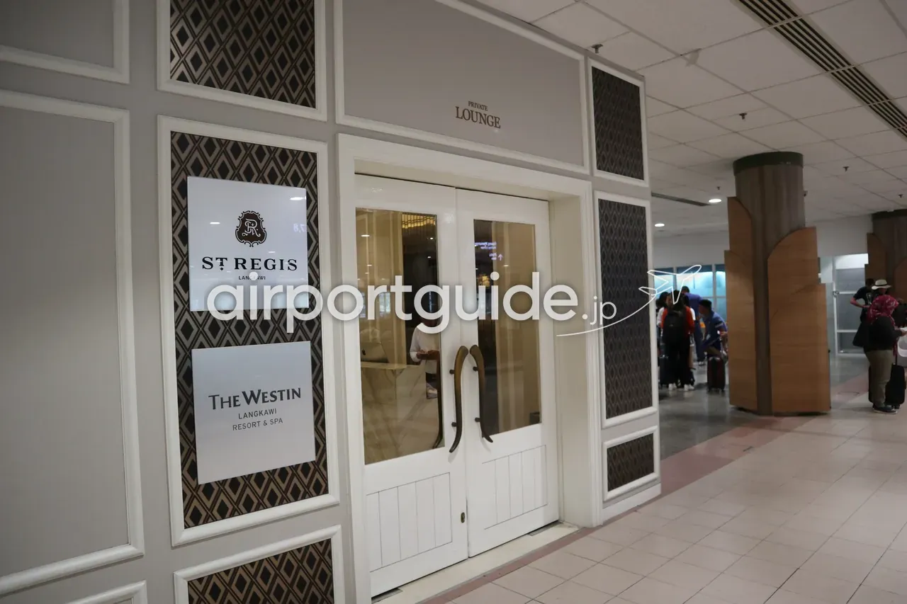 ランカウイ国際空港内の『PRIVATE LOUNGE』入口。白い両開き扉と格子モールディング、ダイヤ柄のパネル、脇に『ST REGIS LANGKAWI』『THE WESTIN LANGKAWI RESORT & SPA』のロゴ看板、右手の廊下に荷物を持つ旅客。