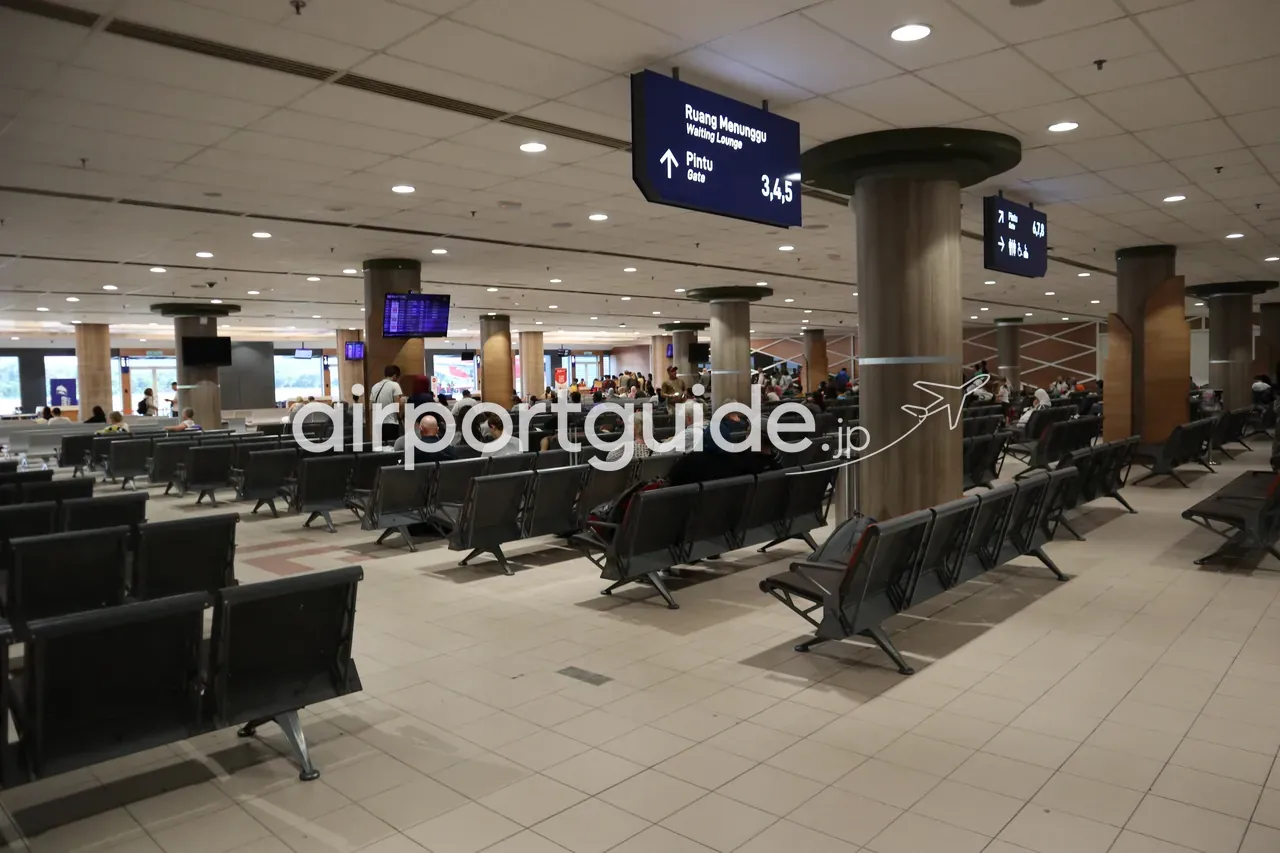 ランカウイ国際空港の搭乗待合エリア。天井から『Ruang Menunggu / Waiting Lounge』『Pintu / Gate 3,4,5』の青い案内板が吊り下がり、グレーの連結ベンチが整然と並び、旅客が待つ。