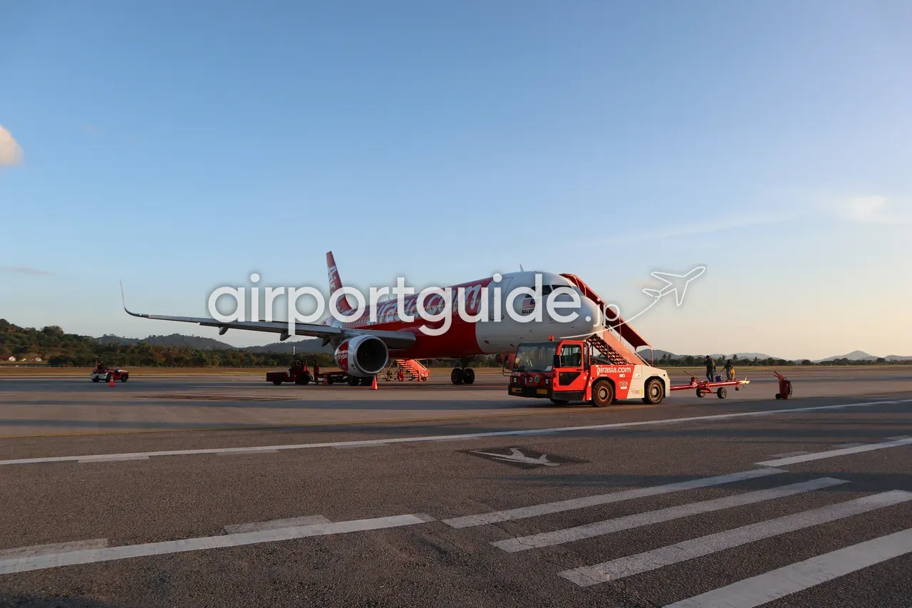 夕方のランカウイ国際空港のエプロンに停まるエアアジア『airasia.com』塗装のA320型機。赤いタラップと『airasia.com』のトーイングカーが付き、背後に島の山並み、金色の夕日が機体を照らす。