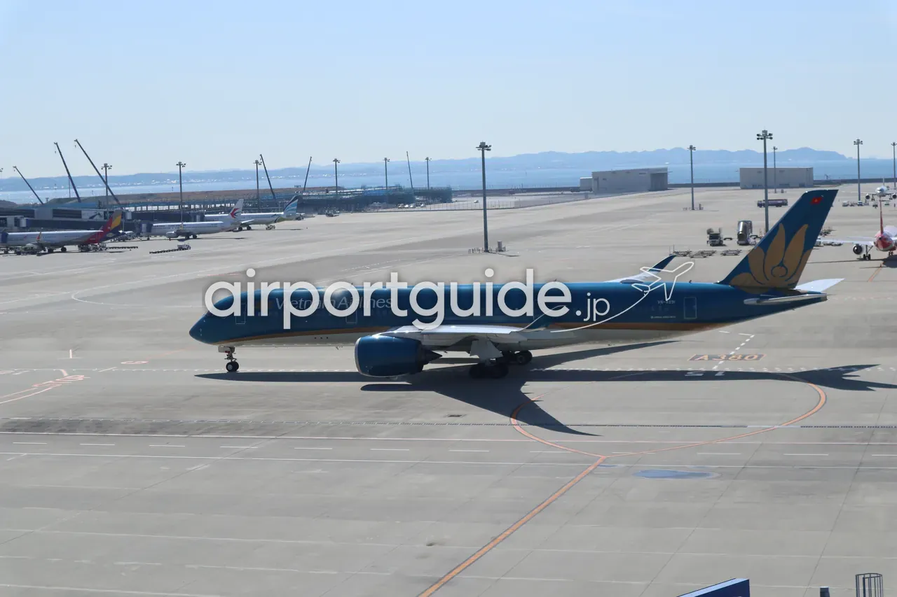 中部国際空港の駐機場を地上走行するベトナム航空のエアバスA350型機。青い胴体に「Vietnam Airlines」、尾翼に黄色い蓮の花のロゴ、エプロンに「A380」のマーキング、背景に他社機と伊勢湾。