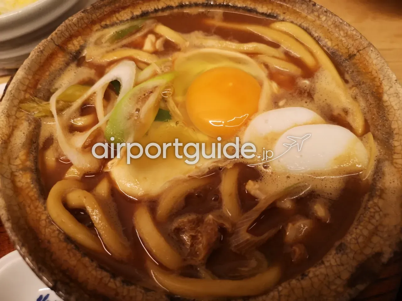 名古屋名物の味噌煮込みうどん。土鍋に濃い味噌だしのうどん、生卵、青ねぎ、かまぼこ、鶏肉が煮込まれ、手前に取り皿と白飯の器。