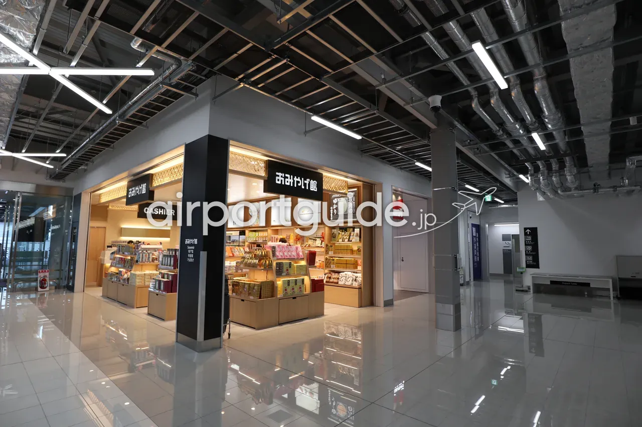 中部国際空港 第2ターミナルの「おみやげ館」土産物店。黒地に白の看板と「CASHIER」表示、木目の陳列棚に箱菓子や地元銘菓が並ぶ。むき出し天井のLCCターミナルらしい内装。