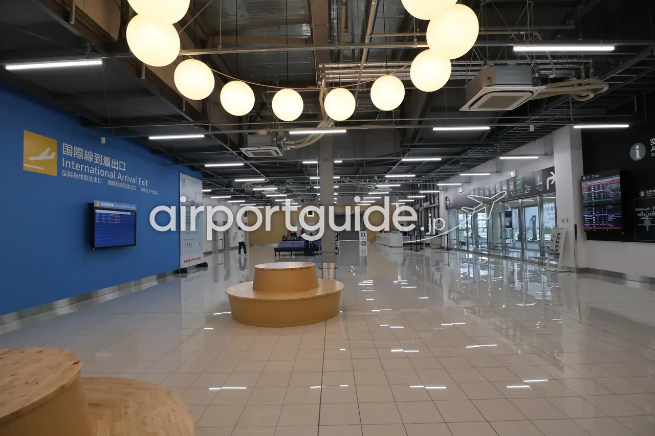 中部国際空港 第2ターミナルの国際線到着出口の広いホール。左壁に「国際線到着出口 International Arrival Exit」の多言語サイン、天井から連なる球形のペンダント照明、木製の円形ベンチ、右手に案内カウンターと出口のガラス扉。