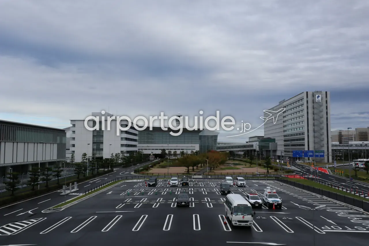 曇り空の中部国際空港を俯瞰した眺め。手前に区画線の引かれた広い駐車場と車道、中央奥にガラス張りのターミナル棟と円筒形の管制塔、右奥に空港直結の高層ホテル。