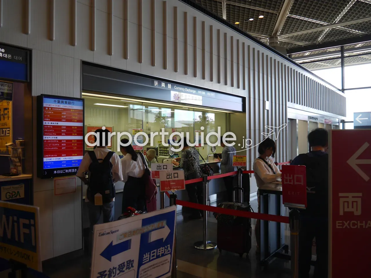 成田空港の外貨両替窓口に旅行者が列を作って順番待ちをしている様子。左手に為替レートの表示ボード、周囲にWiFiレンタルの案内がある。