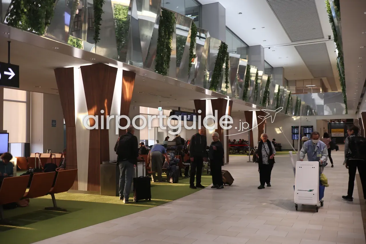 成田空港の制限エリア内コンコース。緑の植栽で装飾された柱と待合席が並び、旅客が行き交う。