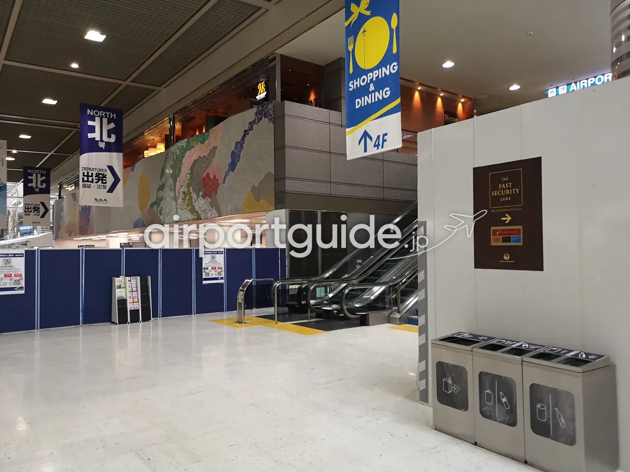 成田空港の案内サインとエスカレーター。「NORTH 北 出発 DEPARTURES」のバナーと「SHOPPING & DINING ↑4F」の吊りサイン、壁面にモザイク壁画。