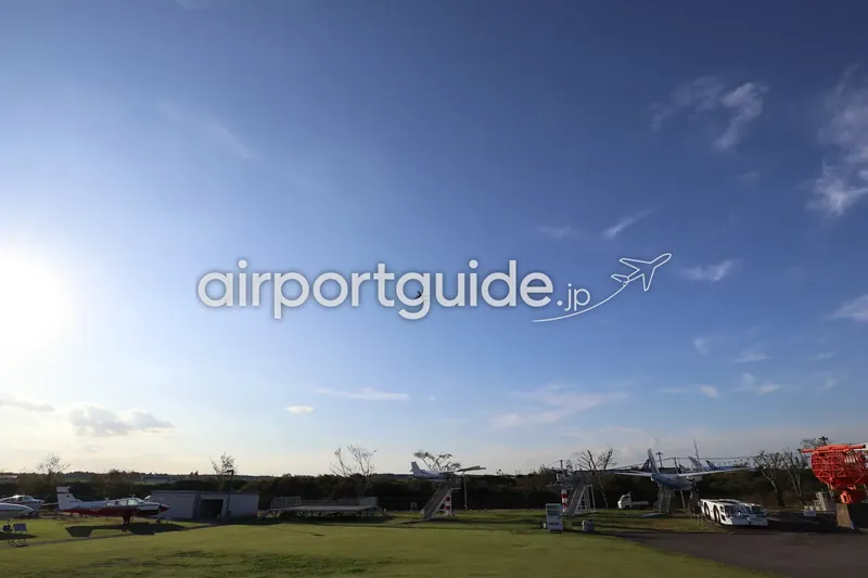 航空科学博物館の屋外展示場の上空を、成田空港を離陸した旅客機が飛んでいく広角の風景。