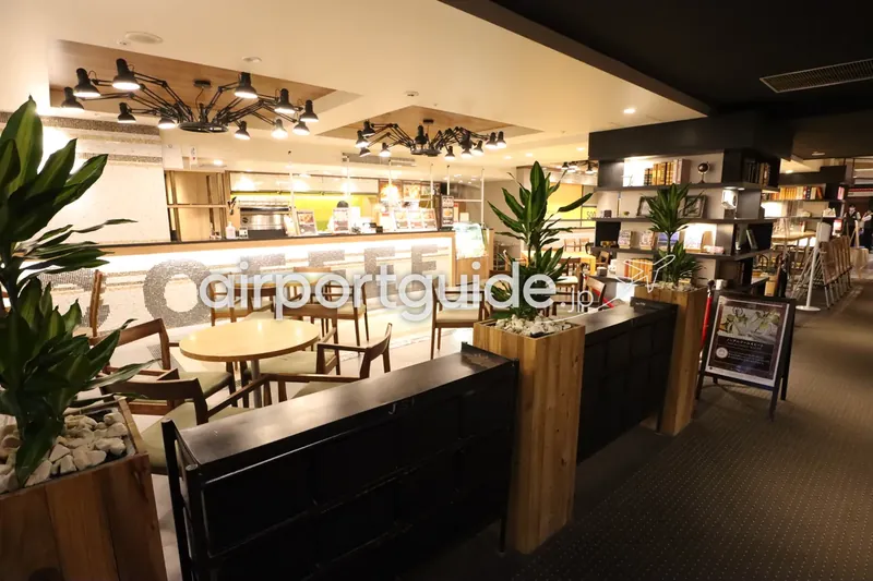 ホテル館内のカフェ。壁の大きな『COFFEE』モザイク文字、木のテーブルと椅子、観葉植物のプランター、奥にカウンターと本棚。