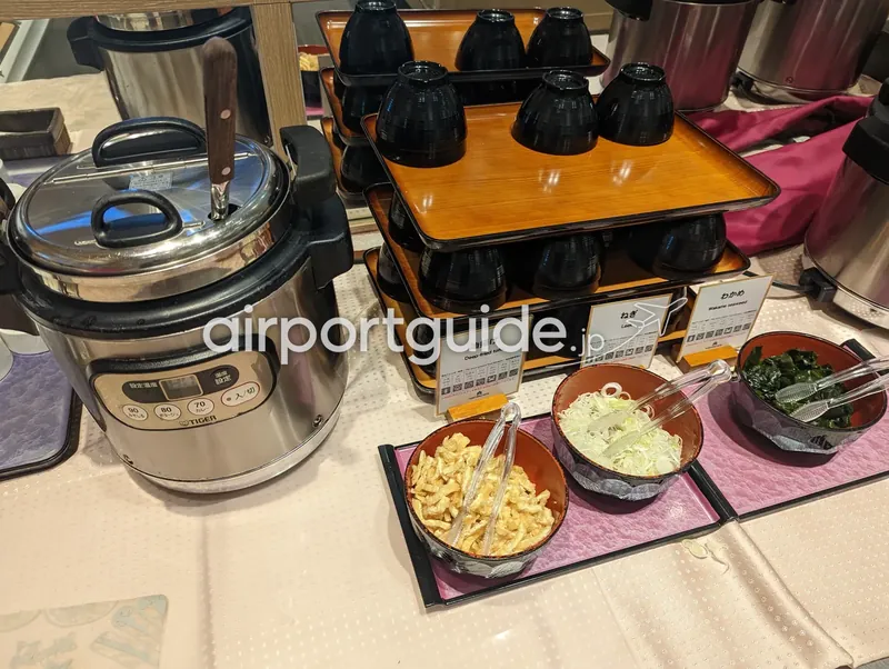 味噌汁・うどんの薬味コーナー。保温ジャーと黒い椀、油揚げ・ねぎ・わかめの薬味が並ぶ。