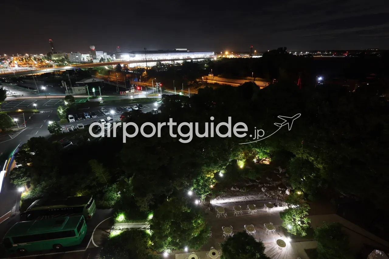 成田東武ホテルエアポートの客室から望む成田空港の夜景。奥にライトアップされたターミナルビル、手前に駐車場やアクセス道路、ホテルの庭園とシャトルバス。