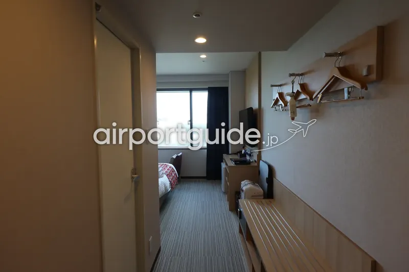 空港近くのホテル客室の入口通路。ハンガー掛けとベンチ、奥に窓とベッド。