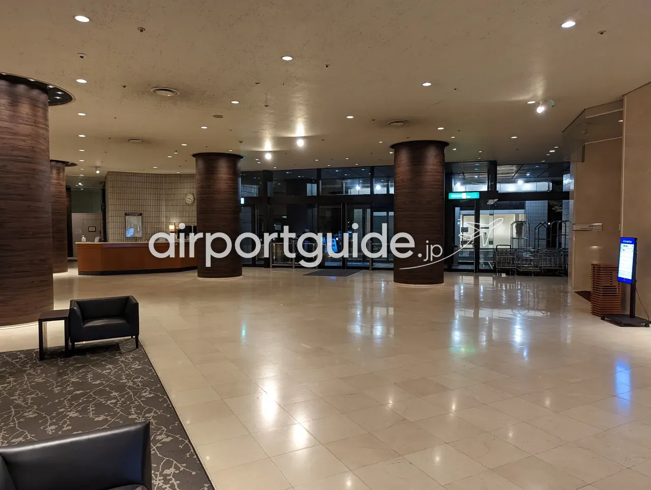 ホテルマイステイズプレミア成田のロビー全景。木目の丸柱、フロントカウンター、自動ドアの入口、荷物カート。