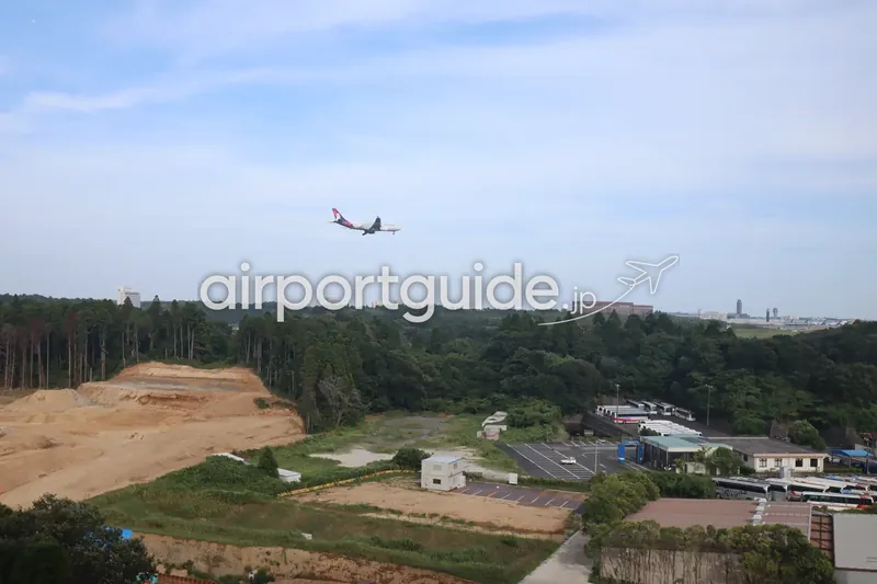 成田空港周辺の森の上空を進入する旅客機。眼下にバス駐車場と造成地。