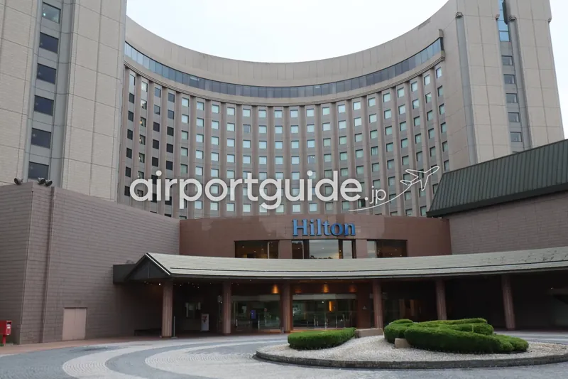 ヒルトン成田の湾曲した客室棟を正面下から見上げた外観。青い「Hilton」ロゴと玄関ポーチ。