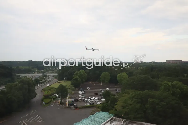 ホテルから見た成田への着陸進入機。白い機体に濃色尾翼の旅客機が郊外上空を降下中。