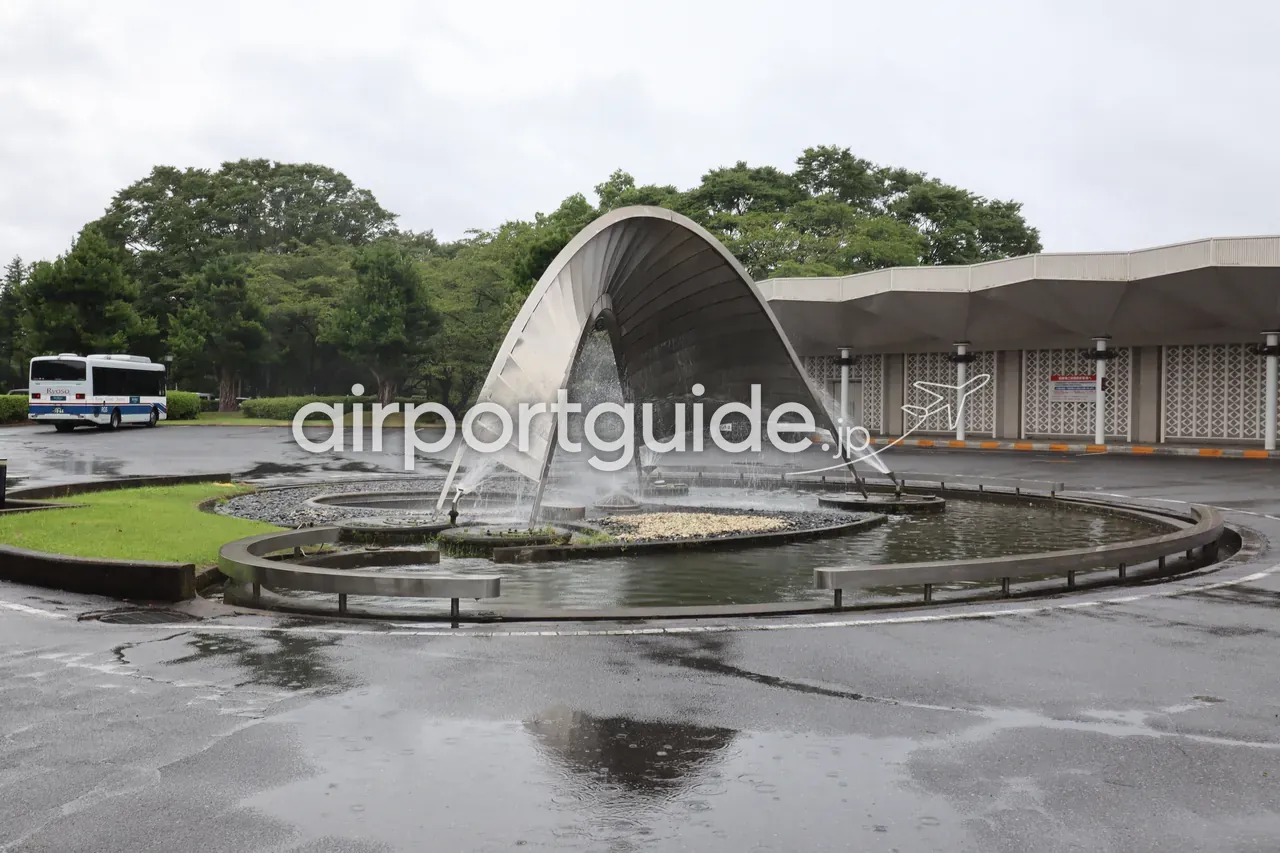 アートホテル成田の車寄せにあるアーチ型の金属製噴水オブジェ。雨で路面が濡れ、左手に送迎バスが停車している。