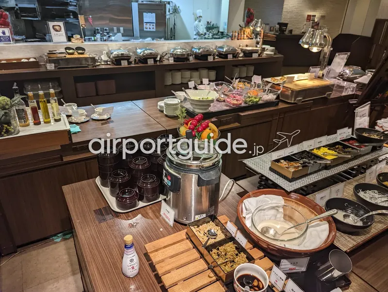 アートホテル成田の朝食レストランのビュッフェ台を斜めから。チェーフィングの列とオープンキッチン、和惣菜や薬味のコーナー。