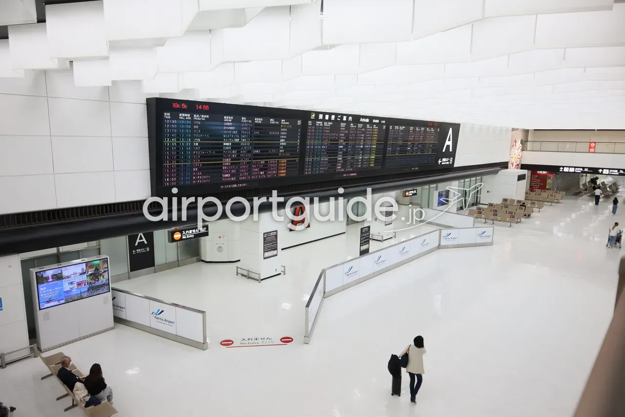 成田空港の到着ロビーを見下ろしたカット。大きな到着便案内板と「出口A」の表示、スーツケースを引いて歩く到着客。