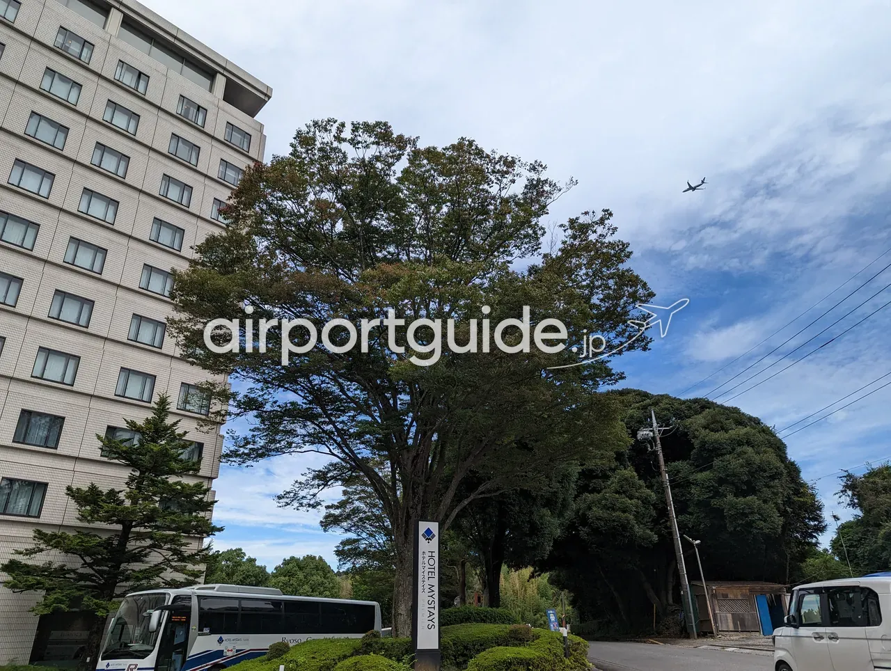 ホテルマイステイズプレミア成田の外観と看板。大きな木の上空を、成田空港へ向かう飛行機が飛んでいる。