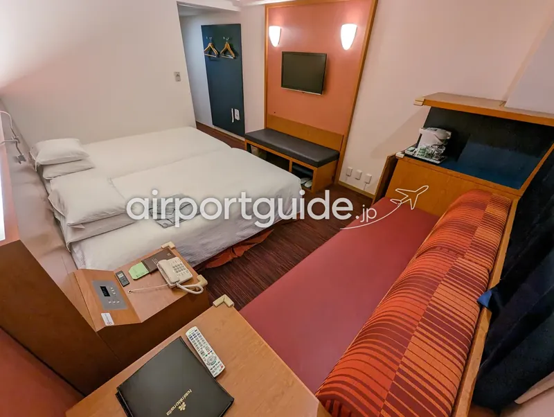 ホテル日航成田の客室を広角で撮影。ベッド脇のテーブルに「hotel Nikko Narita」の案内フォルダとリモコン。