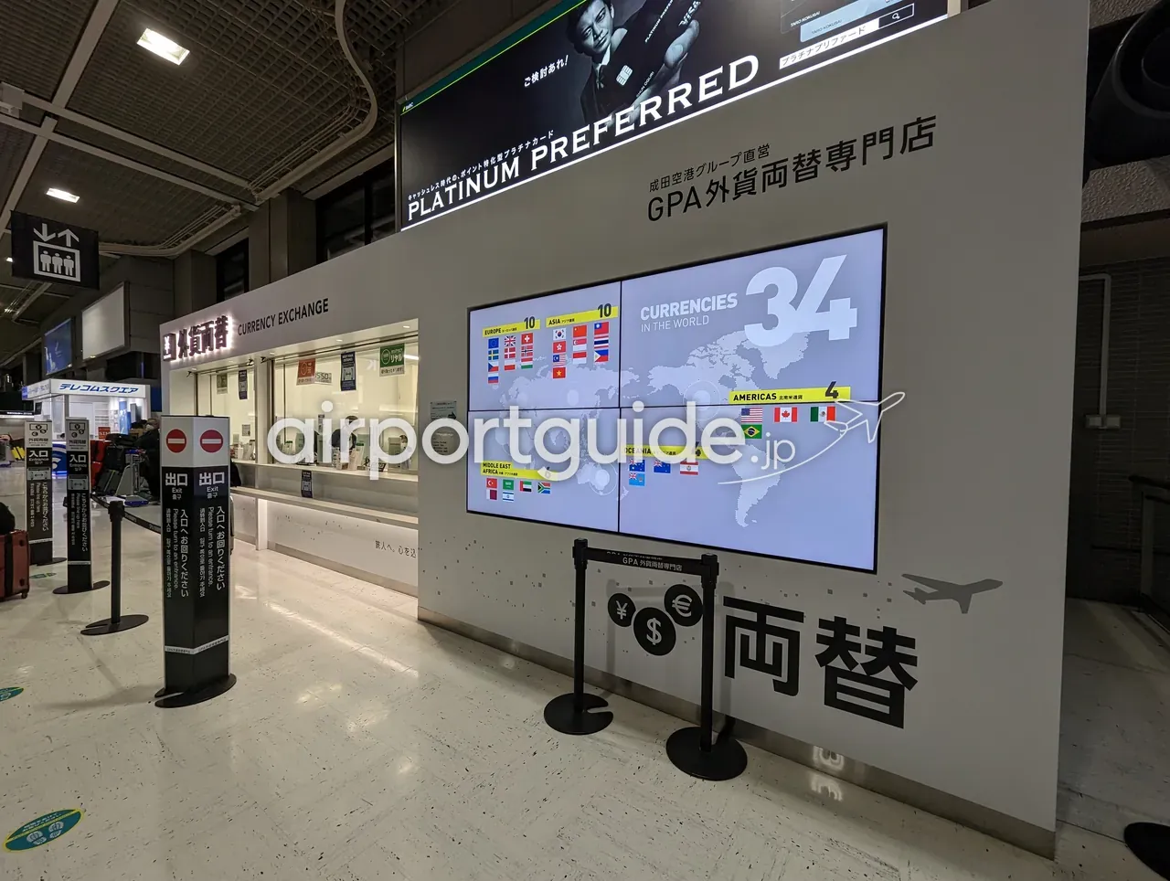 成田空港のGPA外貨両替専門店の窓口と、世界34通貨対応を示す大型デジタルサイネージ。「成田空港グループ直営」の表示と「両替 CURRENCY EXCHANGE」のサイン。
