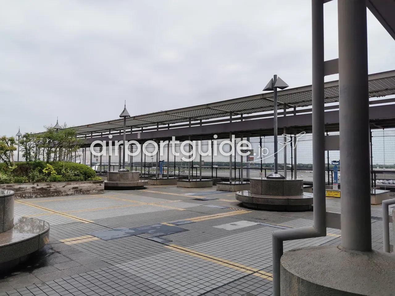成田空港の屋上展望デッキ（送迎デッキ）。屋根付きの通路と金網フェンス越しに、駐機場や滑走路方面を望める屋外スペース。曇天。