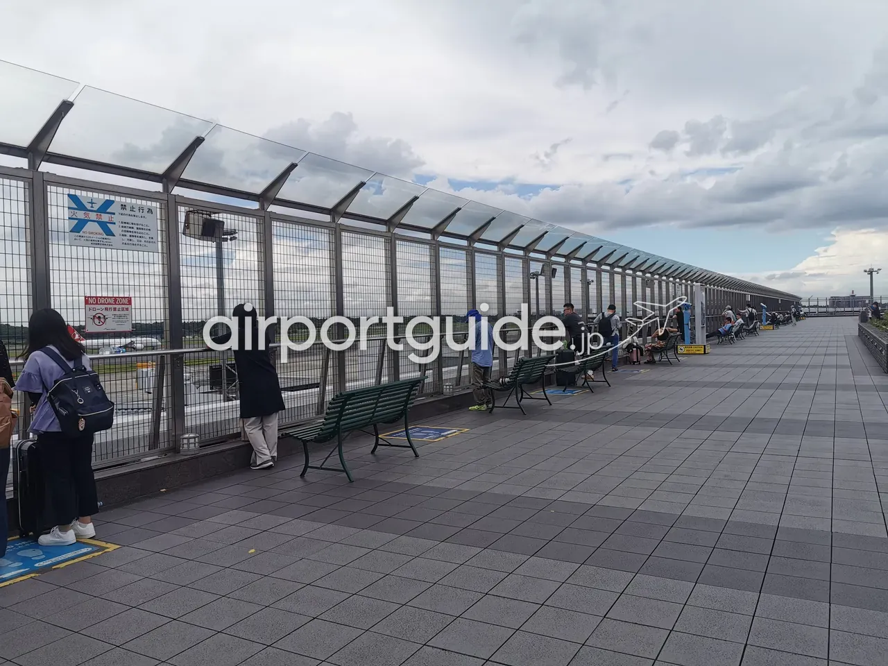 成田空港の屋上展望デッキ。金網フェンス沿いにベンチが並び、旅客が手すりで飛行機を眺める。奥の駐機場に機体が見える。