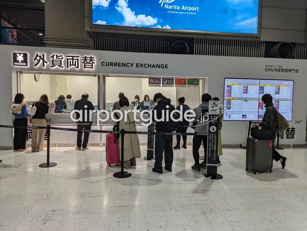 成田空港のGPA外貨両替専門店「外貨両替 CURRENCY EXCHANGE」の窓口に、スーツケースを引いた利用客が列を作って並んでいる様子。右手に大きなデジタルレートボード。