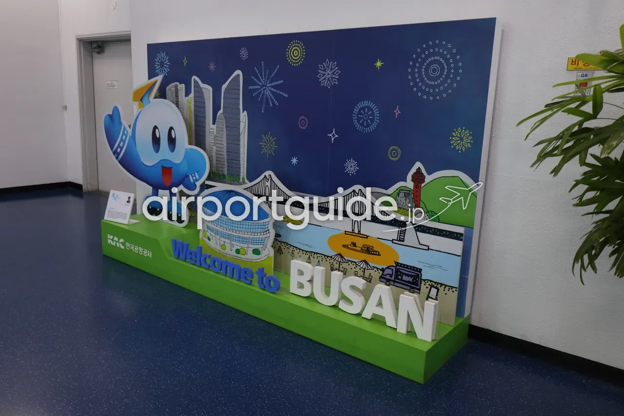 金海空港の到着エリアに置かれた『Welcome to BUSAN』の記念撮影ボード。緑の台に立体文字、青いしずく型のマスコット、広安大橋やBEXCO・海雲台の高層ビル・花火を描いた紺色の背景、左下に韓国空港公社（KAC）のロゴ。