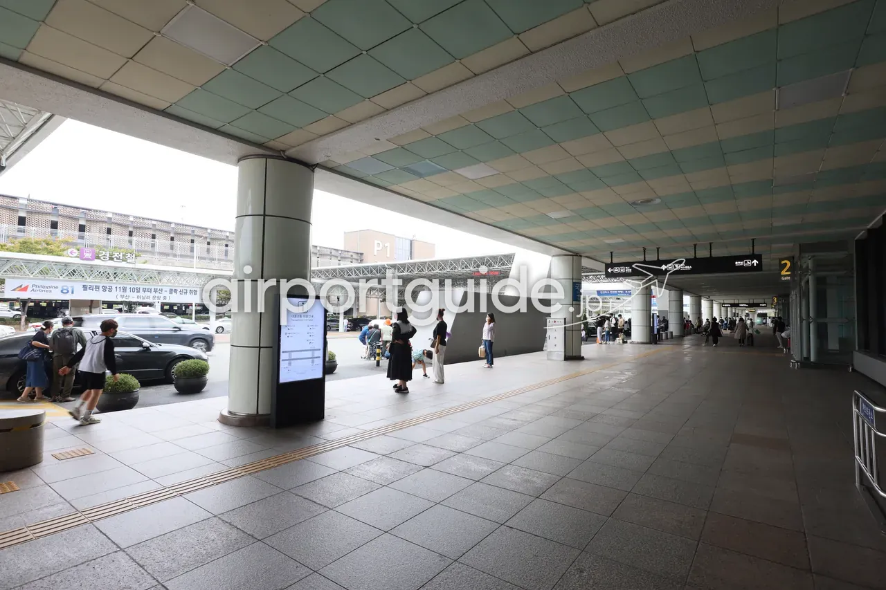 金海空港ターミナル前の屋根付きカーブサイド。柱の奥、道路を挟んだ高架下に『↖ 경전철（軽電車）』の紫の案内サインと駅、フィリピン航空の広告、案内板の前で待つ旅客、右手に『2』のゲート表示。