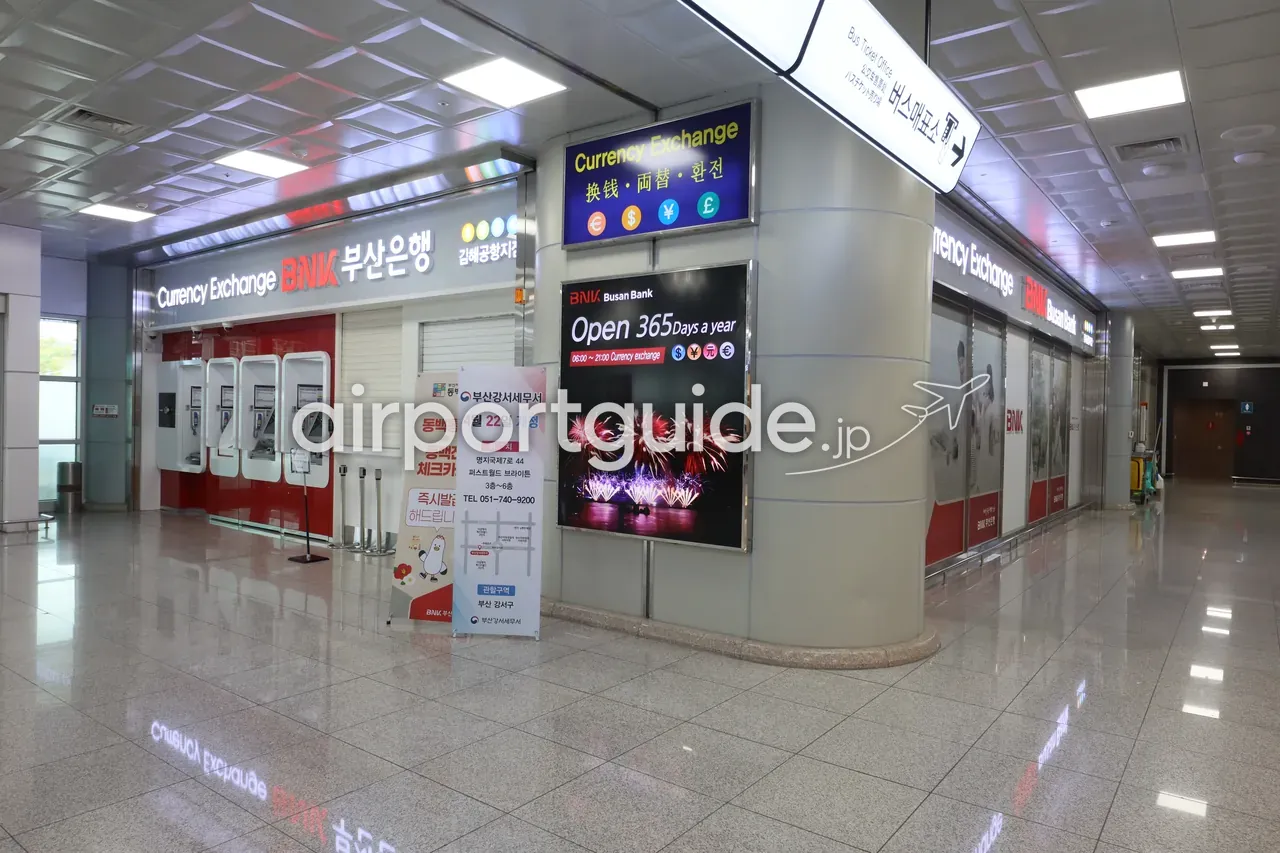 金海空港の両替所「Currency Exchange 换钱・両替・환전」。BNK釜山銀行 金海空港支店のカウンターと無人両替機が並び、「Open 365 Days a year 06:00〜21:00」の案内が出ている。