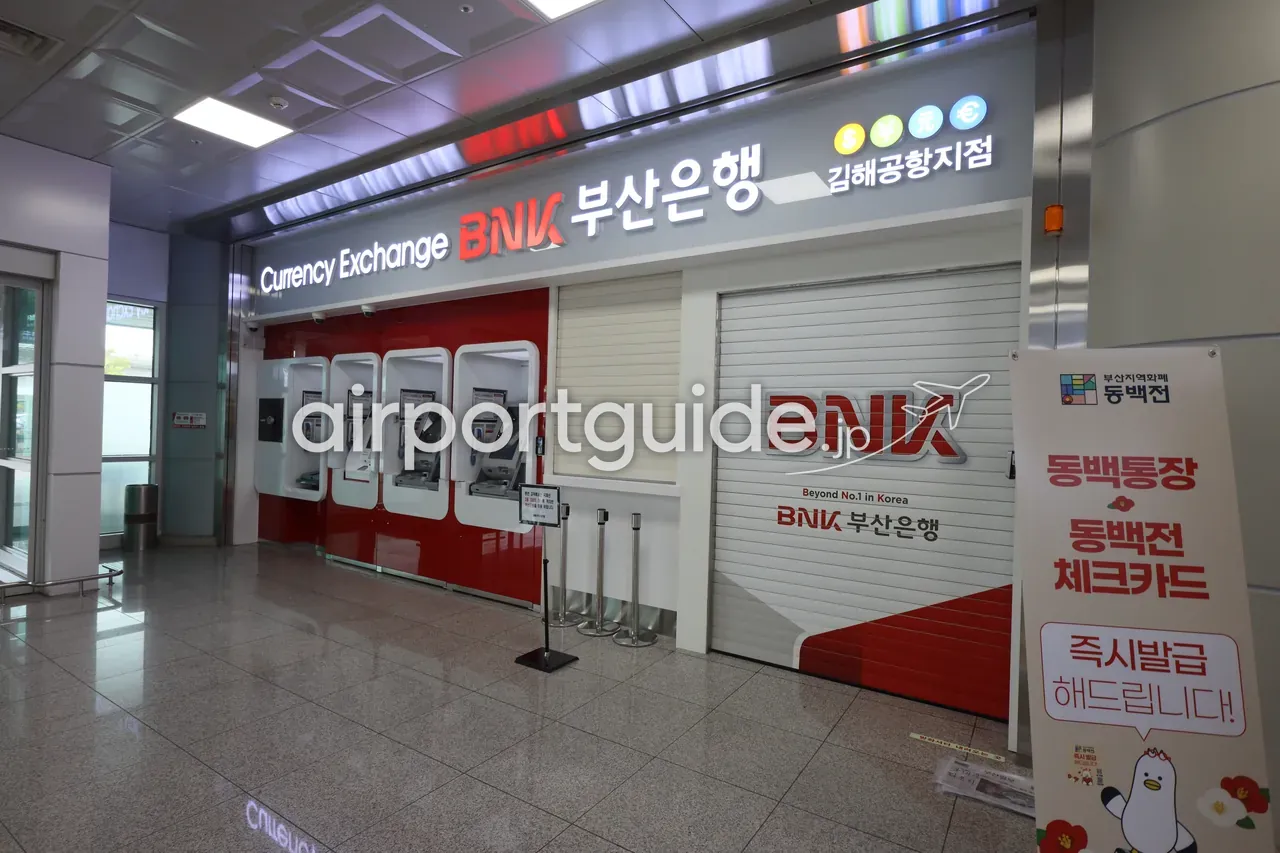 金海空港・国際線到着エリアのBNK釜山銀行 金海空港支店の両替カウンターを正面から。多言語の『Currency Exchange 换钱・両替・환전』看板と、隣に無人両替機（ATM）が並ぶ。