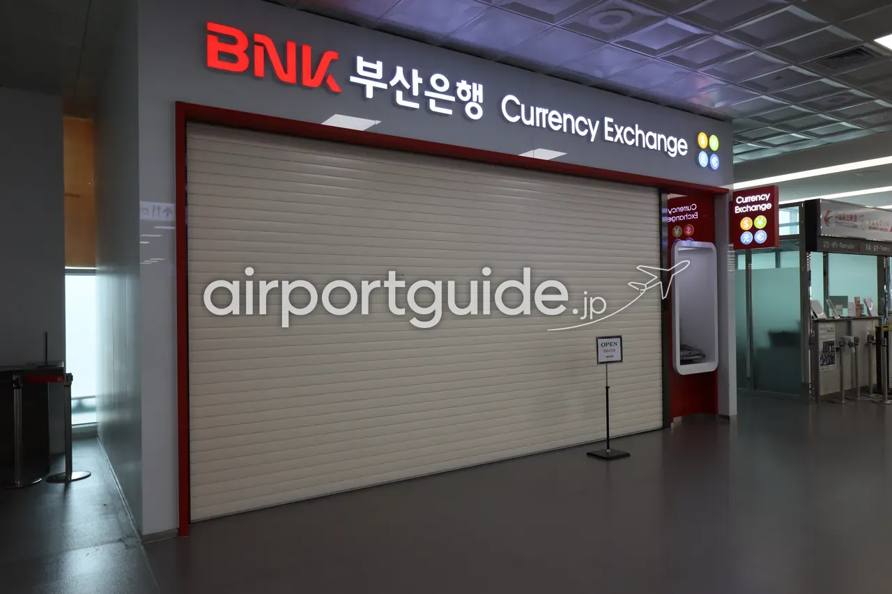 金海空港・国際線到着エリアのBNK釜山銀行の両替ブース。シャッターが下りていて、多言語の『Currency Exchange』看板の下に開店時刻の案内が掲示されている。