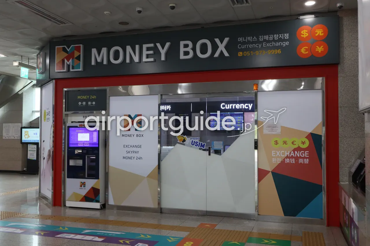 釜山金海軽電車・公港駅の両替店「MONEY BOX（金海空港支店）」。多言語のCurrency Exchange看板、左側に『MONEY 24h 無人両替機』、窓口にはレート板とプリペイドUSIMの案内がある。