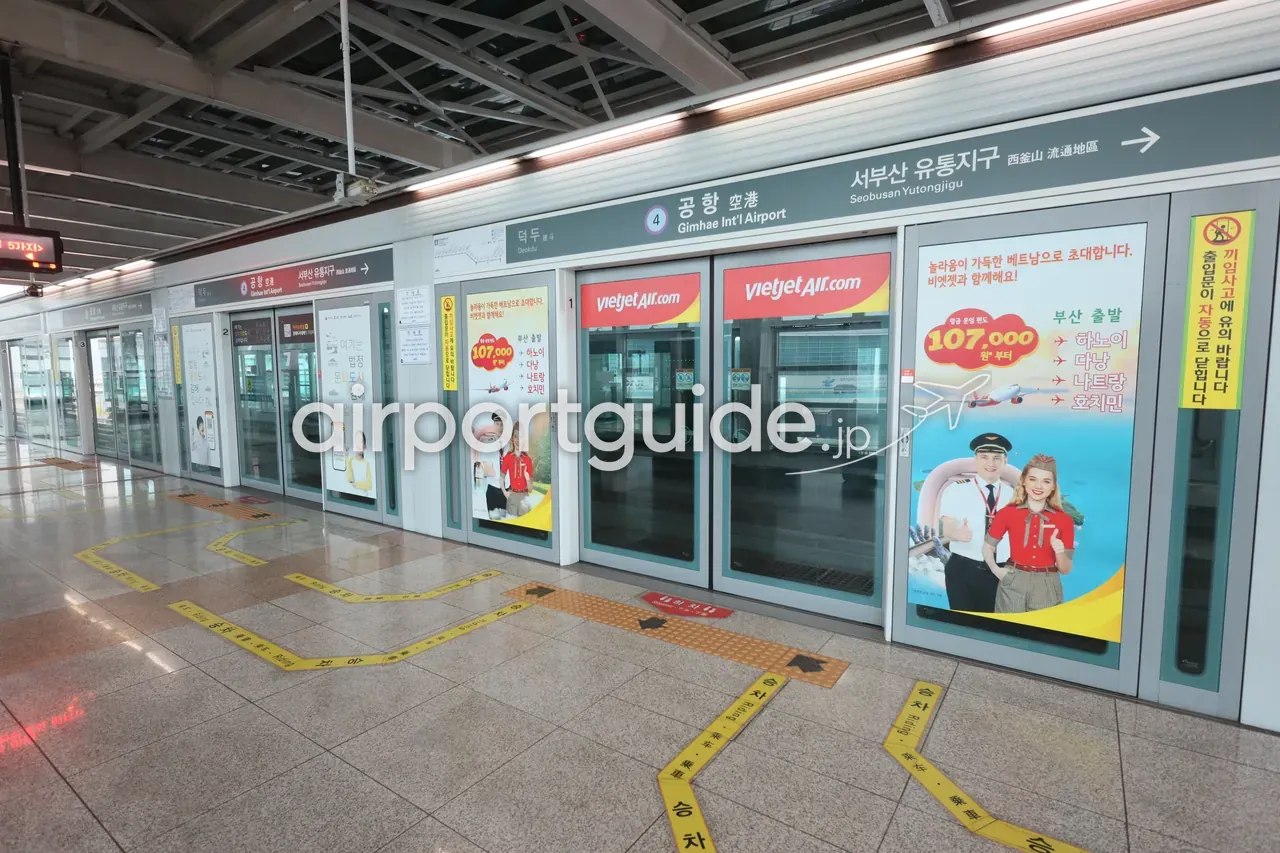 釜山金海軽電車『공항 空港 Gimhae Int'l Airport』駅のホーム。ホームドア（可動式ホーム柵）が並び、次駅『서부산 유통지구 Seobusan Yutongjigu →』の吊り案内、ホームドアにベトナムのビエットジェット航空（VietjetAir.com）の広告。