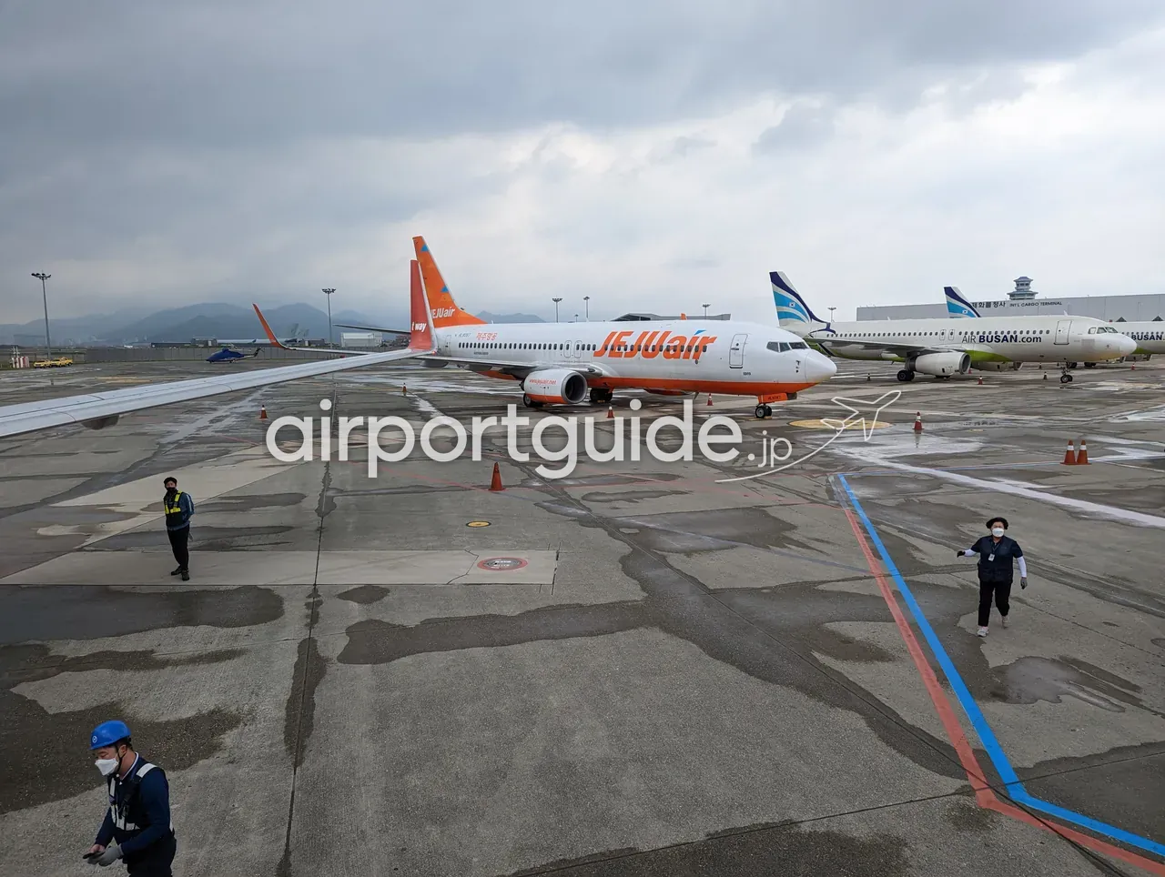 金海空港のエプロン全景。オレンジ塗装のジェジュ航空（JEJUair）のボーイング機、奥にエアプサン（AIR BUSAN.com）のエアバス機、右奥に『INT'L CARGO TERMINAL（国際貨物ターミナル）』の建物と管制塔、地上係員が歩き、遠くに山並み。