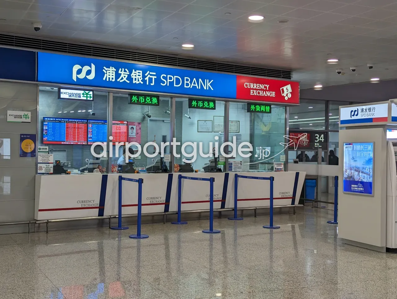 上海浦東空港の両替窓口『浦发银行 SPD BANK／CURRENCY EXCHANGE』。青と赤の看板に『外币兑换』の緑のLED表示とレートボード、白いカウンター、係員が1名、右手に『上海机场 AVINEX』の柱と34番ゲート方面。