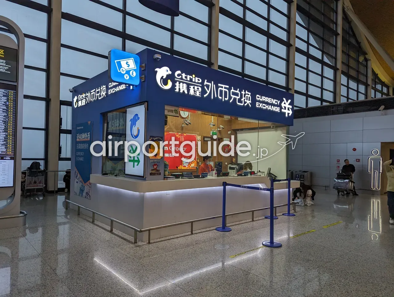 上海浦東空港コンコースの携程（Ctrip）外貨両替ブース。紺色の看板に白いイルカのロゴと「携程 外币兑换 CURRENCY EXCHANGE」、側面に「近40種の外貨・予約不要」の案内。カウンターに係員が1名、背後は大きなガラス窓のコンコース。