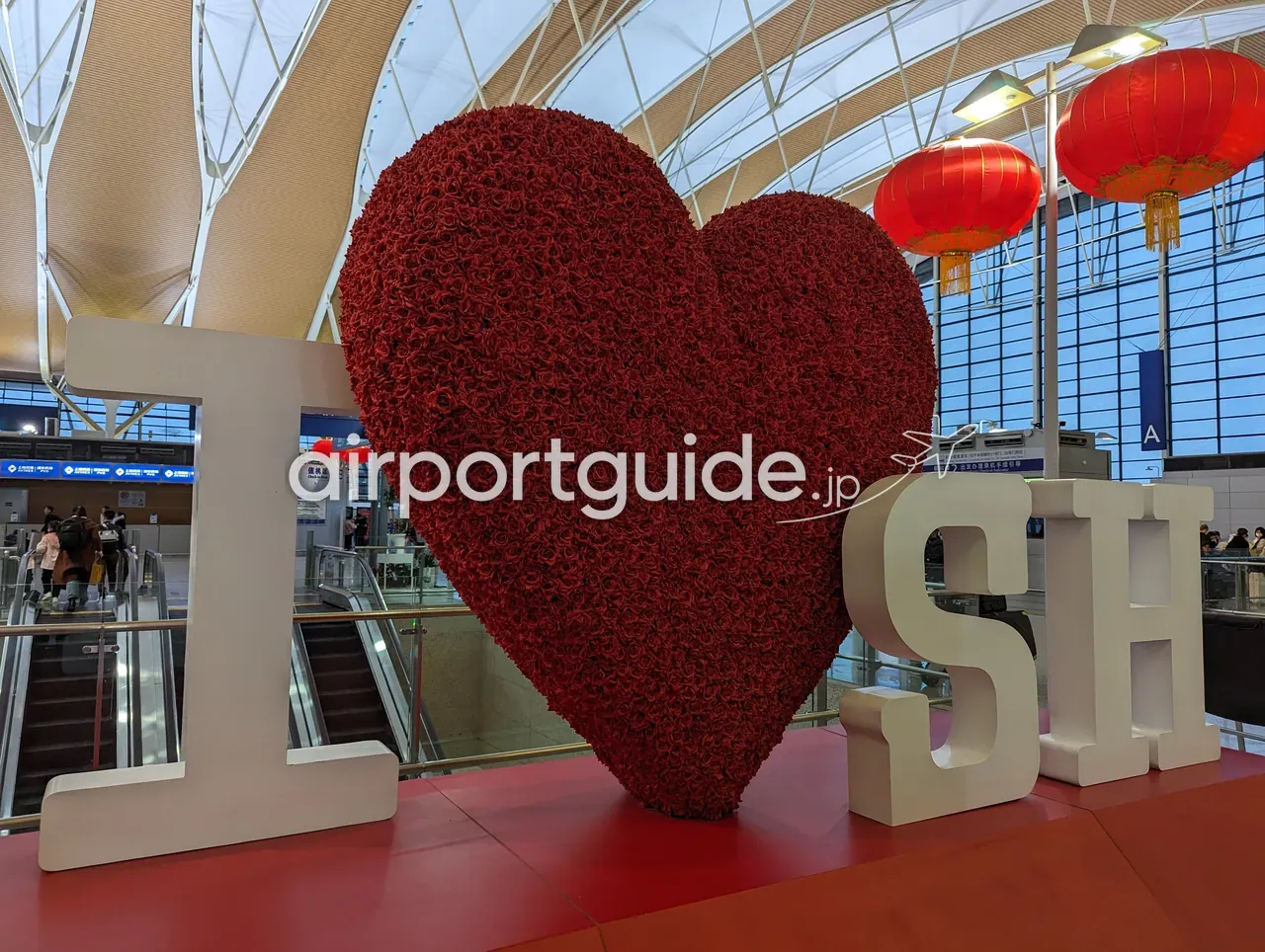 上海浦東空港・出発ホールの『I ♥ SH』(I love Shanghai)フラワーモニュメント。赤いバラで作った大きなハートと白いI・SHの立体文字、波打つ木目の天井、赤提灯、奥にAゲート方面。