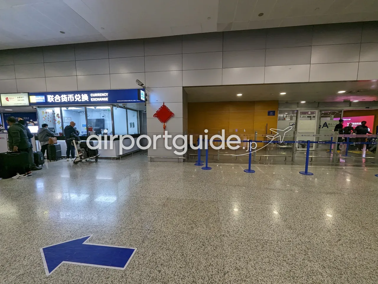 上海浦東空港の到着ホールにある両替所「联合货币兑换 CURRENCY EXCHANGE」。左隣にSIMカード・WiFiの案内、赤い中国結びの飾り。スーツケースやカートを持った到着客が数名、カウンター前にいる。