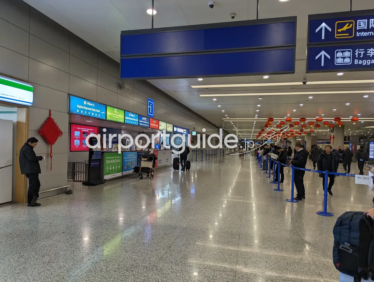上海浦東空港の到着通路。天井に『国际 International』『行李 Baggage』の方向サイン、壁際に『Digital Payment Help Desk／Alipay+』『Information Service』『Convenient Living in SHANGHAI』のカウンター、赤い中国結びと赤提灯、バリア沿いに並ぶ出迎えの人々。