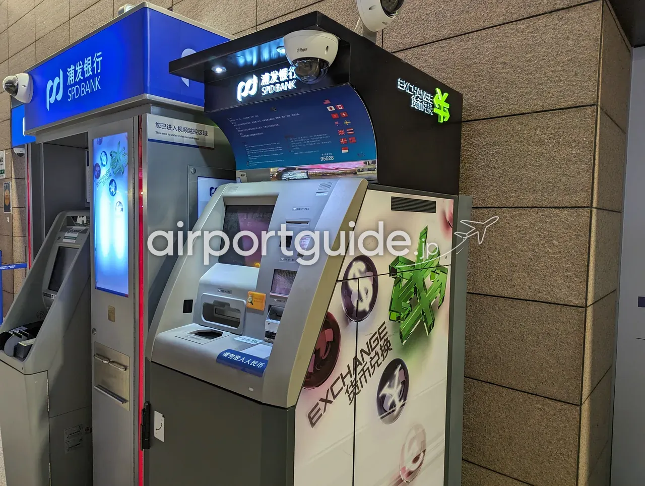 上海浦東空港・浦発銀行（SPD BANK）のATMと、右隣の自動外貨両替機「EXCHANGE 货币兑换」。両替機の画面には各国の国旗が並ぶ。上部にドーム型の防犯カメラと「您已进入视频监控区域 This area is under video surveillance」の表示。