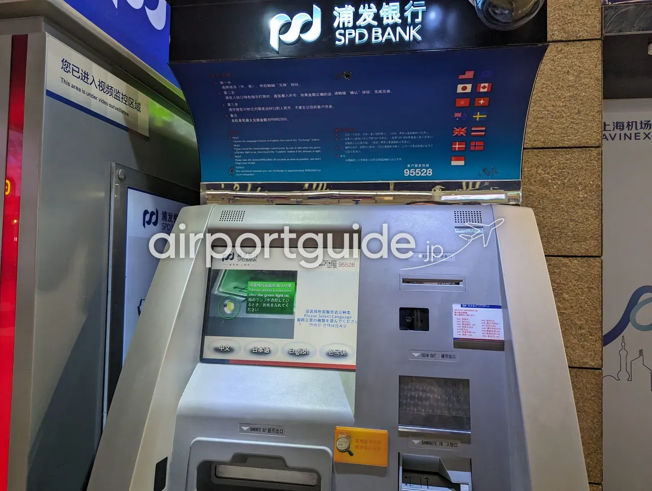 上海浦東空港のSPD BANK自動外貨両替機。操作画面に『请选择所需服务语言种类 Please Select Language 中文／日本語／English／한국어』、上部に各国国旗と手順パネル、『您已进入视频监控区域 This area is under video surveillance』。